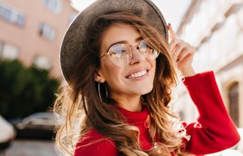 A young woman wearing glasses, a wide-brimmed hat, and a red sweater smiles outdoors on a sunny day.