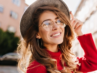 Woman in a red sweater smiles while touching her hat. She is wearing round glasses and hoop earrings, standing outdoors with buildings in the background.