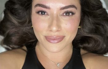 Woman with wavy brown hair lies on her back, wearing a black top and a necklace, looking up and smiling softly at the camera.