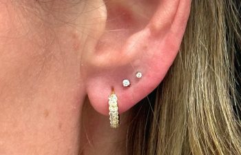 Close-up of an ear with three piercings: a gold hoop earring in the lobe and two small diamond studs in the upper lobe.