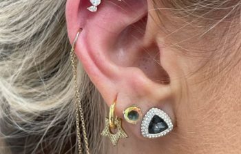 A close-up of an ear with multiple earrings, including a diamond flower stud, a gold hoop with a chain, a star, a green gem, a black triangle, and a teardrop-shaped dangle.