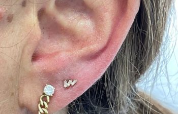 Close-up of a person's ear with two gold earrings: one is a chain design with a small stud, and the other is a small letter "W". Brown hair partially visible.