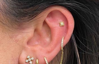 A close-up of an ear with multiple gold and diamond earrings, including a hoop with a chain, studs, and a flower-shaped dangle earring.