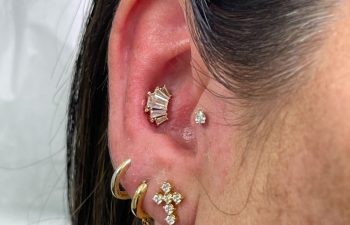 A close-up of a person's ear with multiple gold and diamond earrings, including a fan-shaped stud, a small round stud, and various hoops and a cross-shaped earring.
