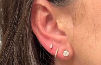 Close-up of a woman's ear with three earrings: a small stud, a short curved bar, and a larger round stud, with brown hair partially visible.
