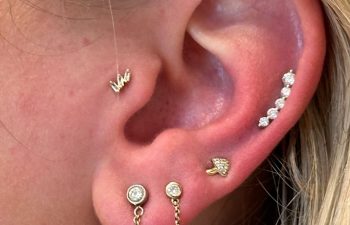 Close-up of a person's ear with multiple piercings, featuring various gold and diamond earrings, including studs, a chain earring, and a curved row of stones.