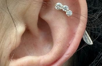 Close-up of an ear with three visible piercings: two small studs and a vertical barbell in the upper cartilage, each adorned with clear gemstones.