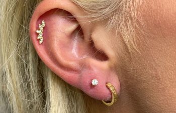 Close-up of a person's ear with three piercings: a gold hoop in the lobe, a small stud above it, and a decorative gold cuff with gemstones on the upper ear.
