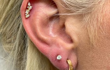 Close-up of a person's ear with three gold and diamond piercings in the cartilage, a diamond stud in the lobe, and a small gold hoop earring below.