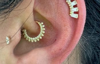 A close-up of an ear with four piercings, each adorned with different gold and diamond earrings, including a crescent-shaped hoop and a rectangular stud cluster.