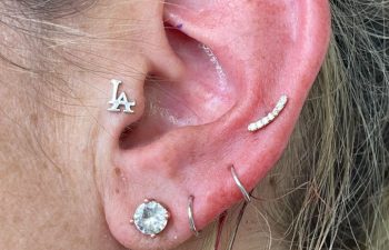 Close-up of a person's ear with multiple piercings, including a hoop, a stud, a jeweled bar, two rings, and an "LA" letter earring.