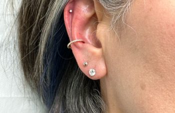 Close-up of a person's ear with short gray hair, showing four piercings with various earrings: a stud in the lobe, a small hoop and stud in the helix, and another small stud above the lobe.