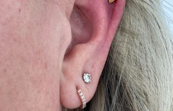 Close-up of a person's ear with three earrings: one small gold stud in the upper ear, one clear stud in the earlobe, and one small hoop with stones in the lower lobe.