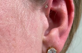 Close-up of a person's ear with two piercings: a small gold stud in the cartilage and a larger gold stud with a clear stone in the earlobe.