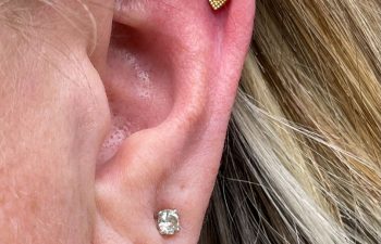 Close-up of an ear with three piercings: a small hoop in the lobe, a round stud above it, and a gold teardrop-shaped stud with a central gem on the upper cartilage.