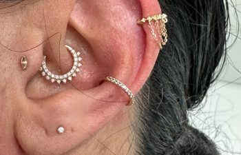 A close-up of an ear with multiple piercings, featuring gold and diamond jewelry including a hoop with small gems, a bar with chains, and several studs.