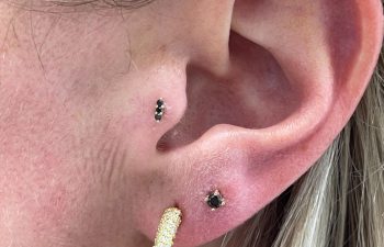 Close-up of a person's ear with three earrings: a gold textured hoop in the lobe, a small black stud above it, and another small black stud in the upper ear cartilage.