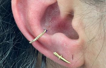Close-up of an ear with two gold earrings: a small hoop in the cartilage and a dagger-shaped stud in the earlobe.