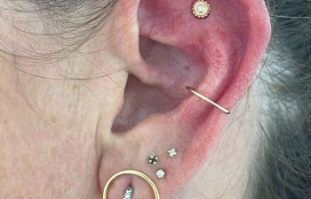 Close-up of an ear with multiple piercings, including gold hoops and studs with small clear stones and a larger circular gold earring with embedded stones in the lobe.