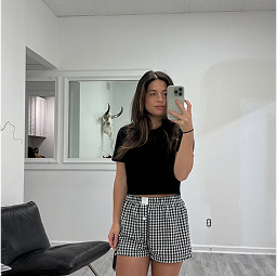 A woman stands in a modern, white room taking a mirror selfie. She wears a black t-shirt and checkered shorts. A black chair and a wall-mounted animal skull are visible in the background.