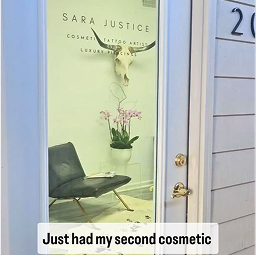 A glass door with "Sara Justice Cosmetic Tattoo Artist Luxury Studio" written on it; inside, a chair, orchid, and wall-mounted skull are visible.