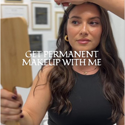A woman with long brown hair holds a handheld mirror, looking at her reflection. Text over the image reads: “GET PERMANENT MAKEUP WITH ME.”.