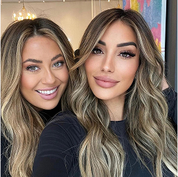 Two women with long, wavy, highlighted hair and wearing black tops smile for a selfie indoors.