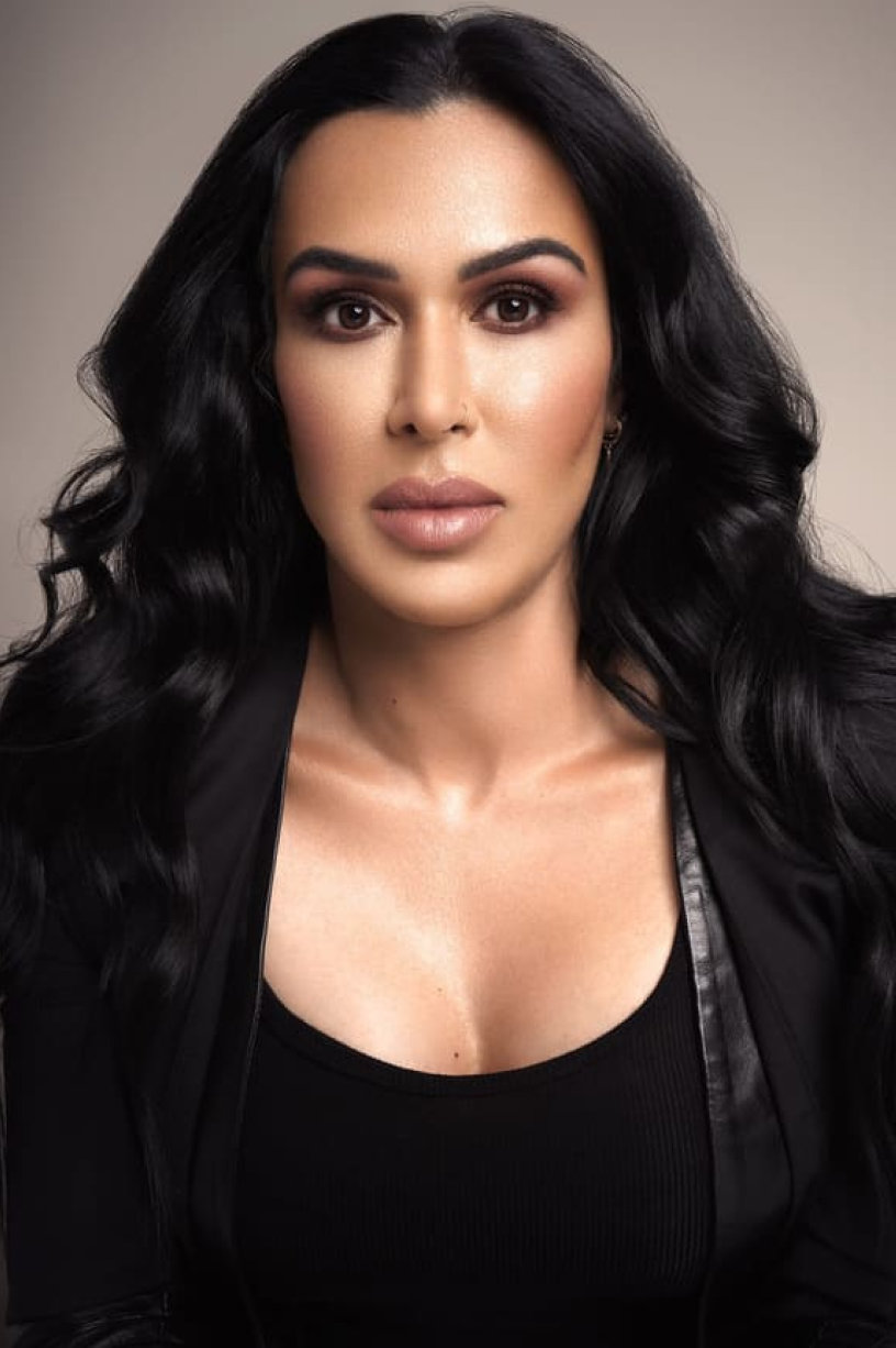 A person with long, wavy black hair and natural makeup, wearing a black top and black jacket, poses against a neutral background.