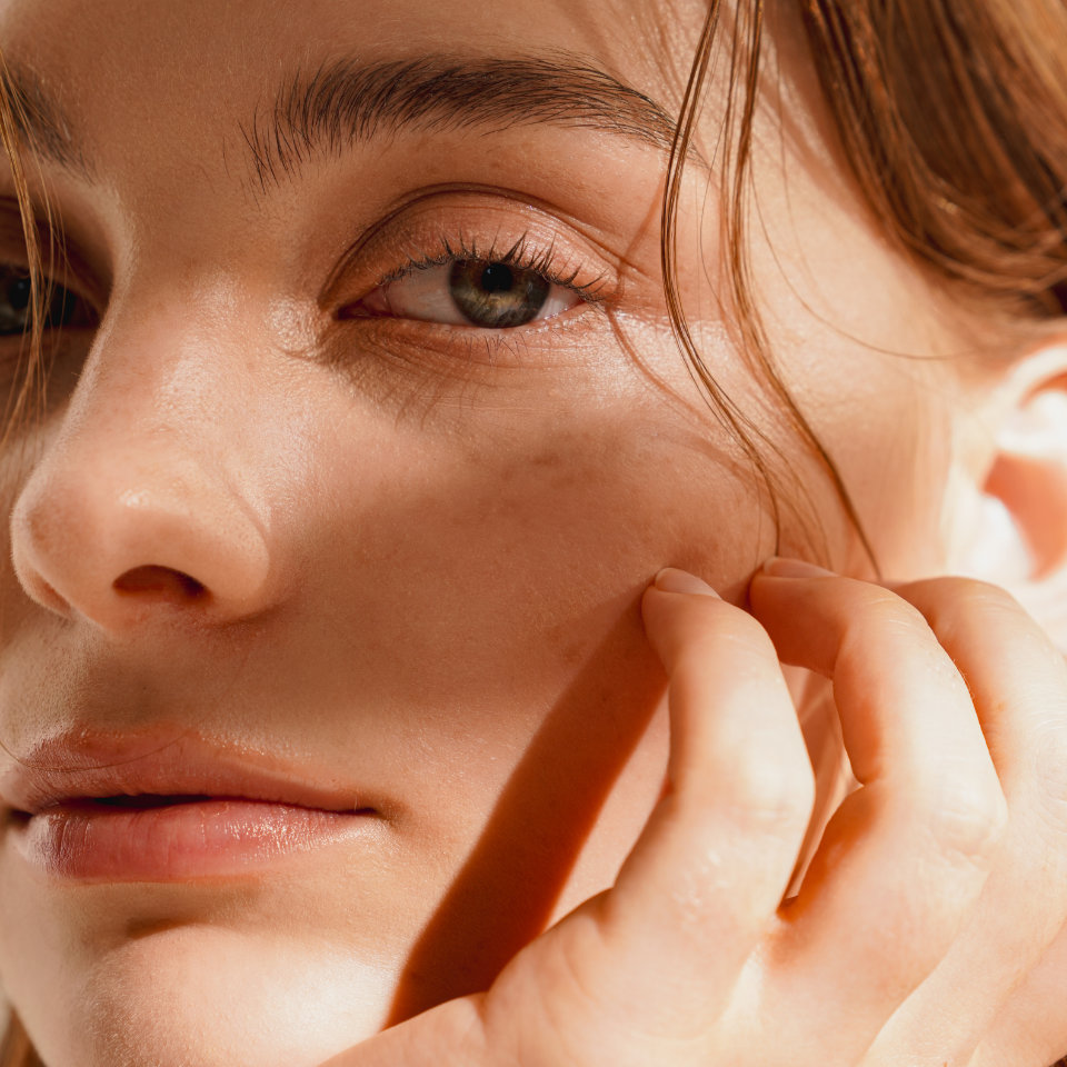 Close-up of a person's face with light brown hair, showing clear skin, subtle freckles, and a hand gently touching their cheek in natural daylight.