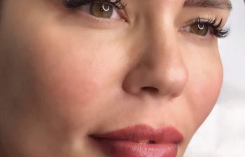 Close-up of a woman's face with well-defined eyebrows, long eyelashes, smooth skin, and red lipstick, looking slightly to the side.
