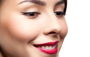 Close-up of a woman's face showing smooth skin, red lipstick, winged eyeliner, and neatly shaped eyebrows on a white background.