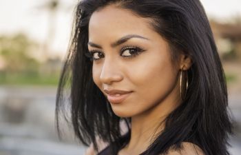 Woman with straight dark hair and gold hoop earrings looks at the camera outdoors, smiling slightly.