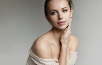 A young woman with light skin and blue eyes poses against a neutral background, wearing an off-shoulder beige sweater and gently touching her face with one hand.
