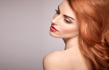 Woman with long red hair and red lipstick looks over her shoulder against a neutral background.