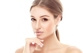 A woman with light brown hair tied back, clear skin, and natural makeup rests her chin on her hand against a white background.