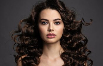 Woman with long, voluminous, dark curly hair and natural makeup poses against a plain dark background, looking directly at the camera.