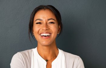 A woman with dark hair pulled back smiles at the camera, wearing a light sweater over a white top, standing against a dark gray background.