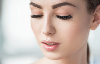 A close-up of a woman's face with her eyes closed, showing smooth skin, thick eyebrows, and long, dark eyelashes.