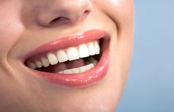 Close-up of a person's mouth smiling, showing white teeth and glossy lips against a pale blue background.