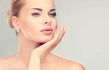 A woman with clear skin and light eyes rests her chin on her hand, looking slightly to the side against a plain background.