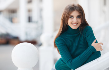 A woman with long brown hair wearing a teal turtleneck sweater leans against a white railing and smiles at the camera.