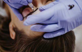 A person wearing purple gloves uses a handheld tool to apply pigment to another person's eyebrow, likely performing a cosmetic tattoo procedure.