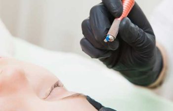 A gloved hand holds a microblading tool near a person's eyebrow, preparing for a cosmetic procedure.