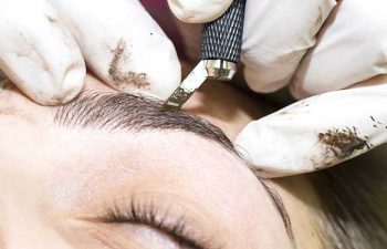 A close-up of a person’s eyebrow being microbladed by a gloved technician using a handheld tool.