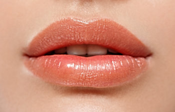 Close-up of a person's lips with a natural peach-colored lipstick, slightly parted, showing a small gap between the upper and lower teeth.
