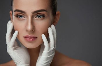 A woman with clear skin is gently touching her face with both hands while wearing white medical gloves, against a plain grey background.