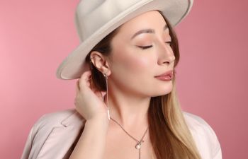 Woman wearing a white hat and light-colored blazer poses against a pink background with her hand touching her ear and eyes closed.