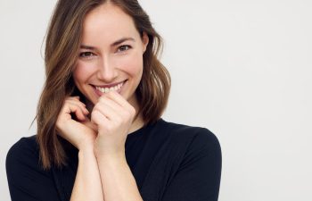 Woman with shoulder-length brown hair smiles at the camera, hands held near her face, wearing a black long-sleeve top against a plain light background.