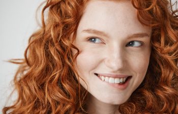 Close-up of a young person with curly red hair and fair skin, smiling and looking slightly to the side against a neutral background.