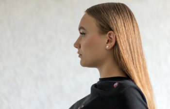 A woman with straight, light brown hair is shown in profile, wearing a black top and pearl earrings, against a light, plain background.